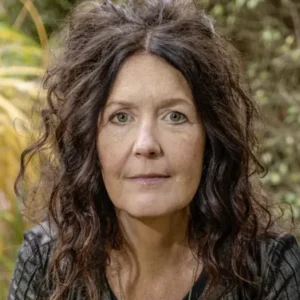 Close-up portrait of a woman with dark, wavy hair outdoors, wearing a dark top and a neutral expression; blurred garden background.